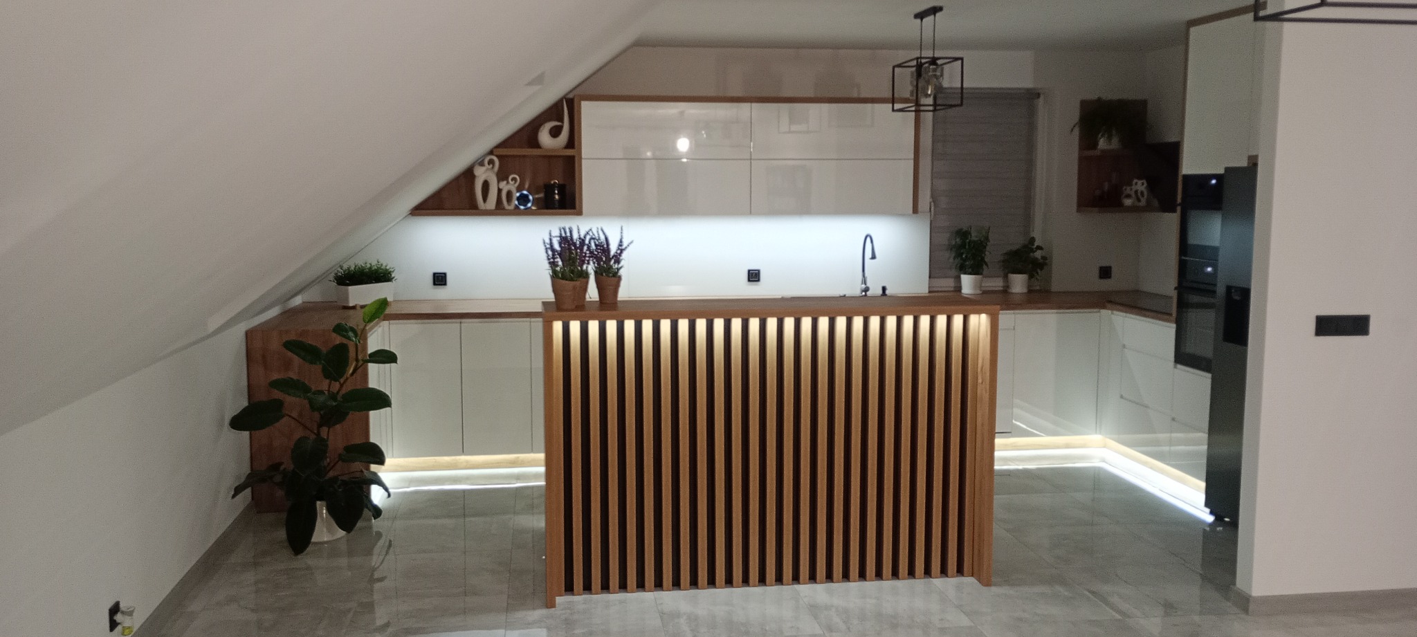 Nowoczesna kuchnia w skosach z drewnianym barkiem z podświetleniem LED, białe szafki i drewniane blaty. Uwagę przykuwa geometryczna lampa i rośliny doniczkowe.