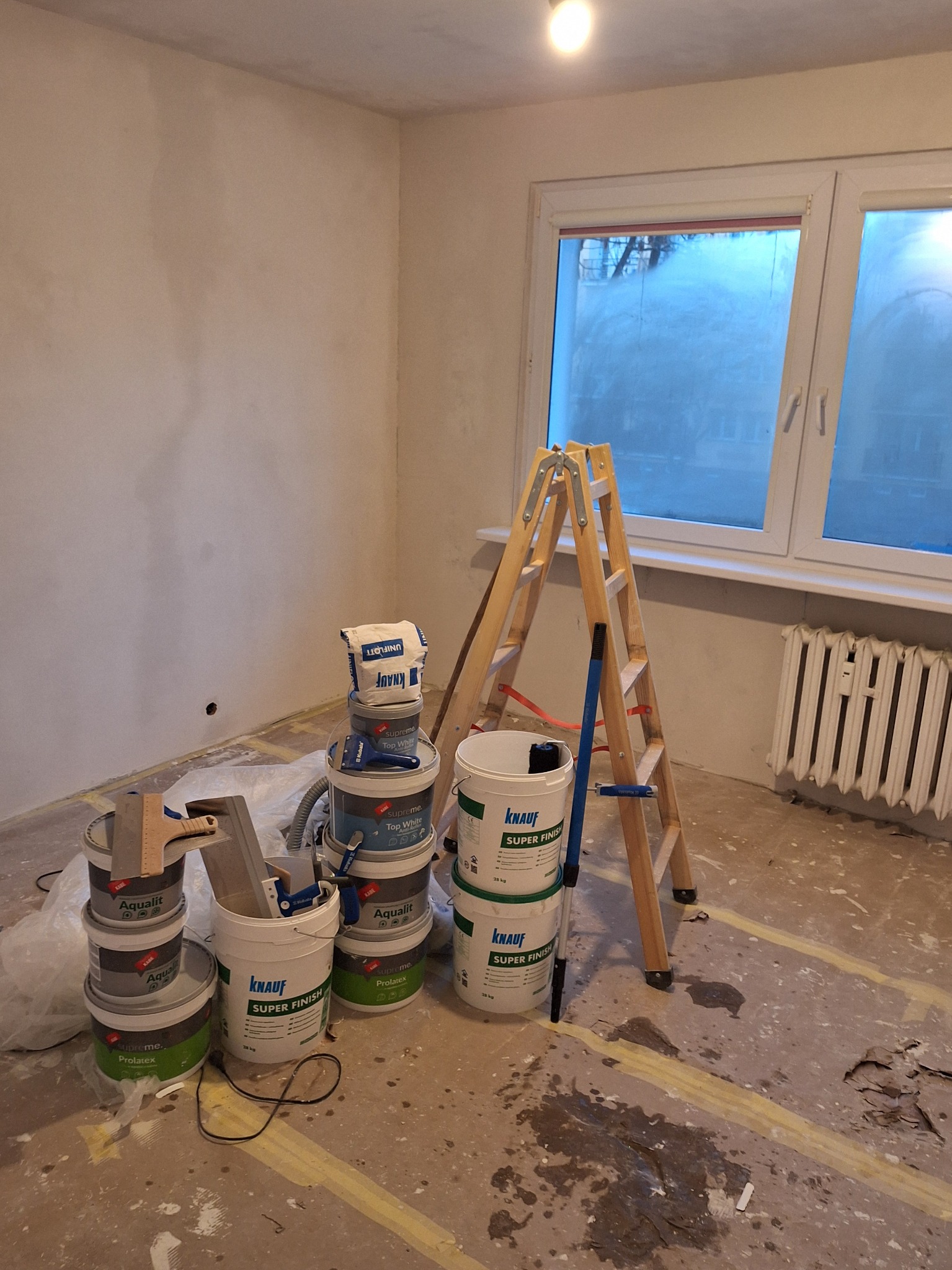 Pokój w trakcie prac wykończeniowych: ściany po gładzi, drabina, farby Knauf i Aqualit, zabezpieczona podłoga. Widok na okno z zamgloną szybą.