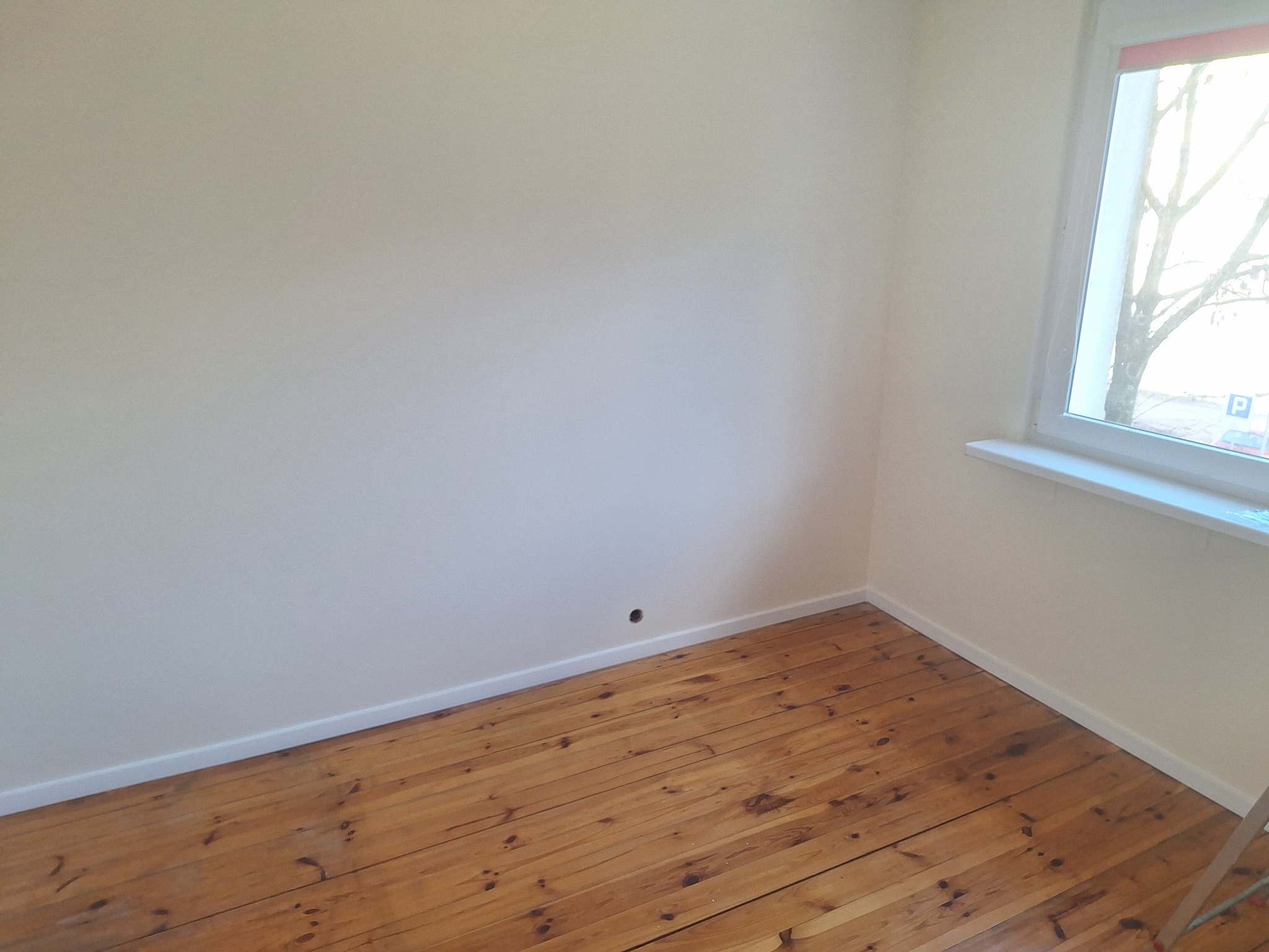 Jasny pokój po odświeżeniu: kremowe ściany, drewniana podłoga, białe listwy przypodłogowe i okno z widokiem na drzewo. Minimalistyczne wykończenie wnętrza.