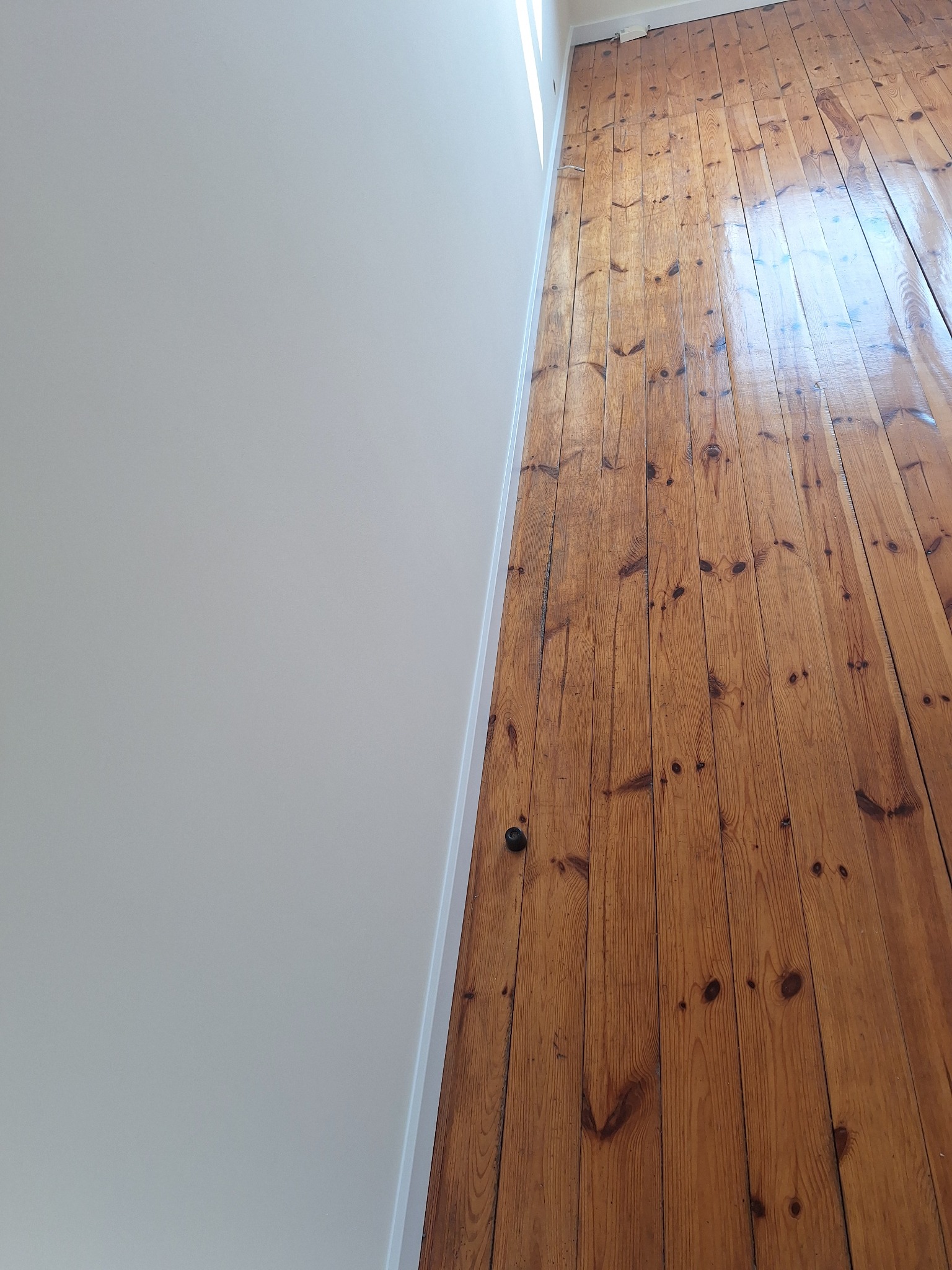 Jasna, świeżo pomalowana ściana sąsiaduje z odnowioną, lakierowaną podłogą z desek. Widoczne detale drewna i gładka powierzchnia ściany.