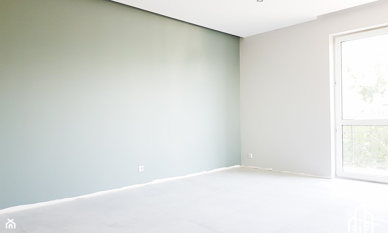Jasne wnętrze po malowaniu: ściana w odcieniach zieleni i szarości, biała podłoga zabezpieczona folią, okno z widokiem na zieleń. Minimalistyczny design, świeżo po remoncie.