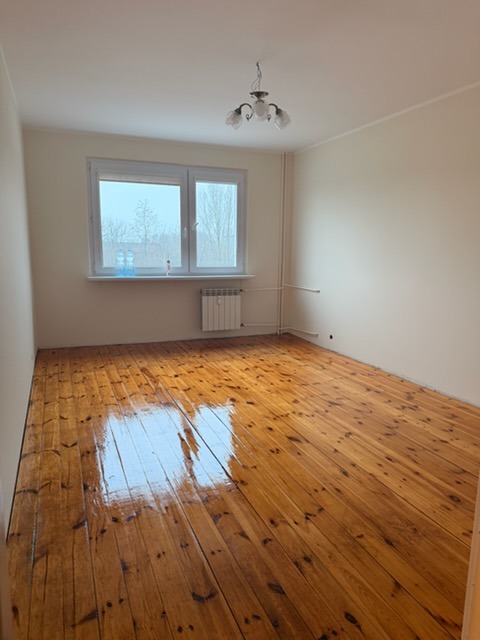 Pomalowane na kremowo puste pomieszczenie z oknem i błyszczącą, lakierowaną drewnianą podłogą. Widoczny grzejnik i lampa sufitowa.