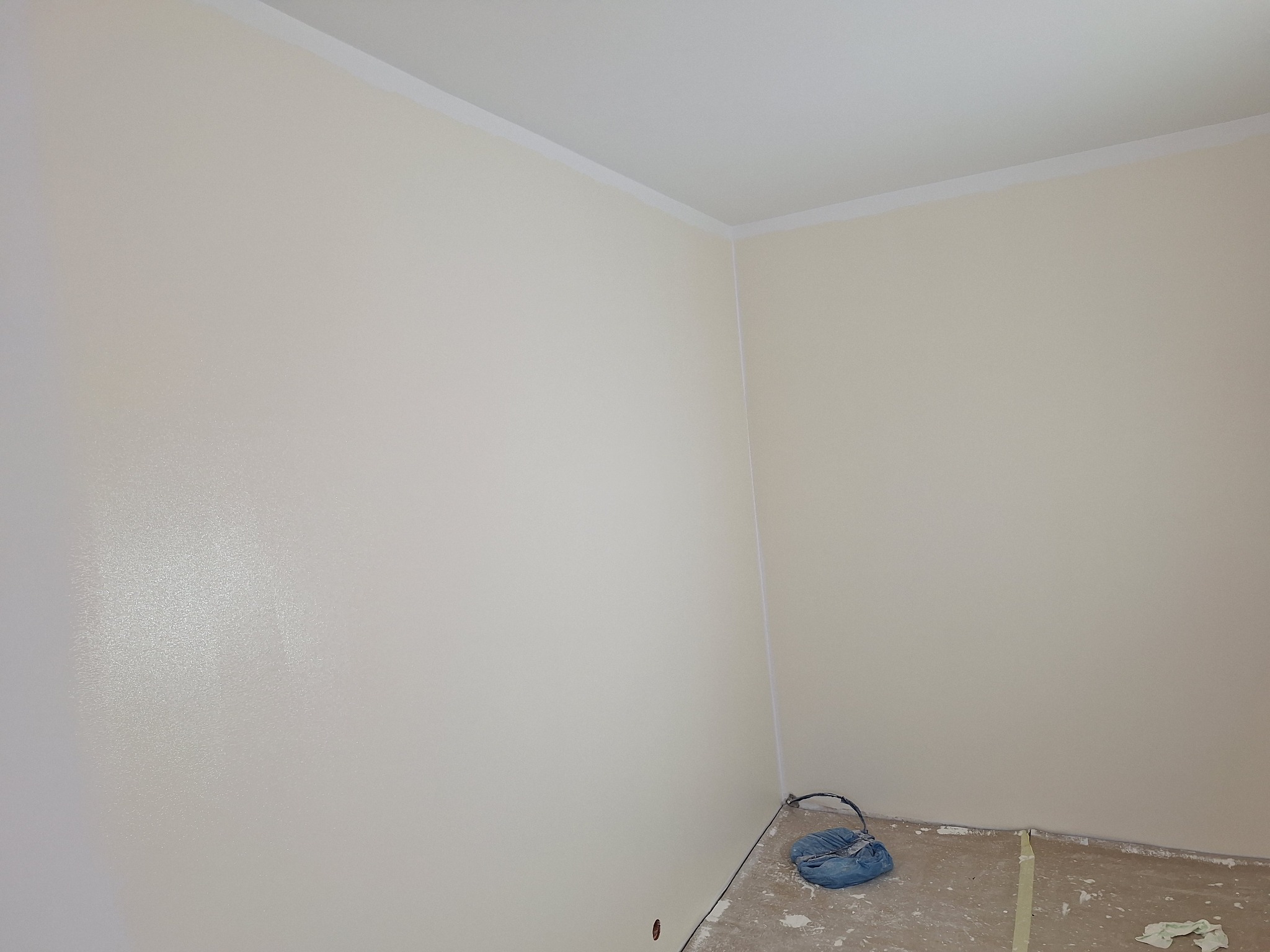 Świeżo pomalowane, kremowe ściany w rogu pokoju, z widocznym workiem malarskim na podłodze i taśmą malarską przy listwach. Gładka powierzchnia, jasne oświetlenie.