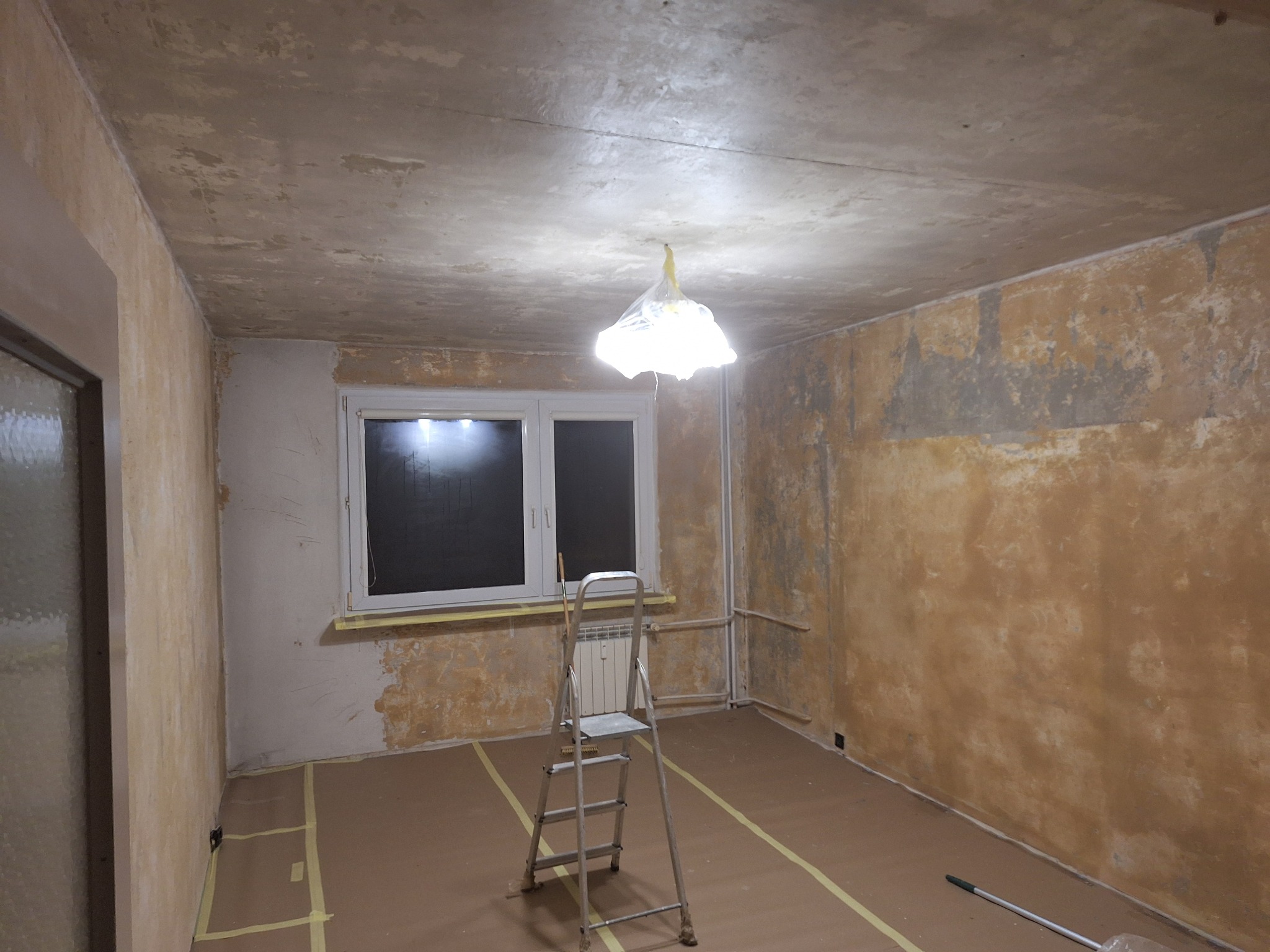Pokój w trakcie remontu: ściany z resztkami starej farby, zabezpieczona podłoga, drabina i zakryta lampa. Przygotowanie do gładzi lub malowania.