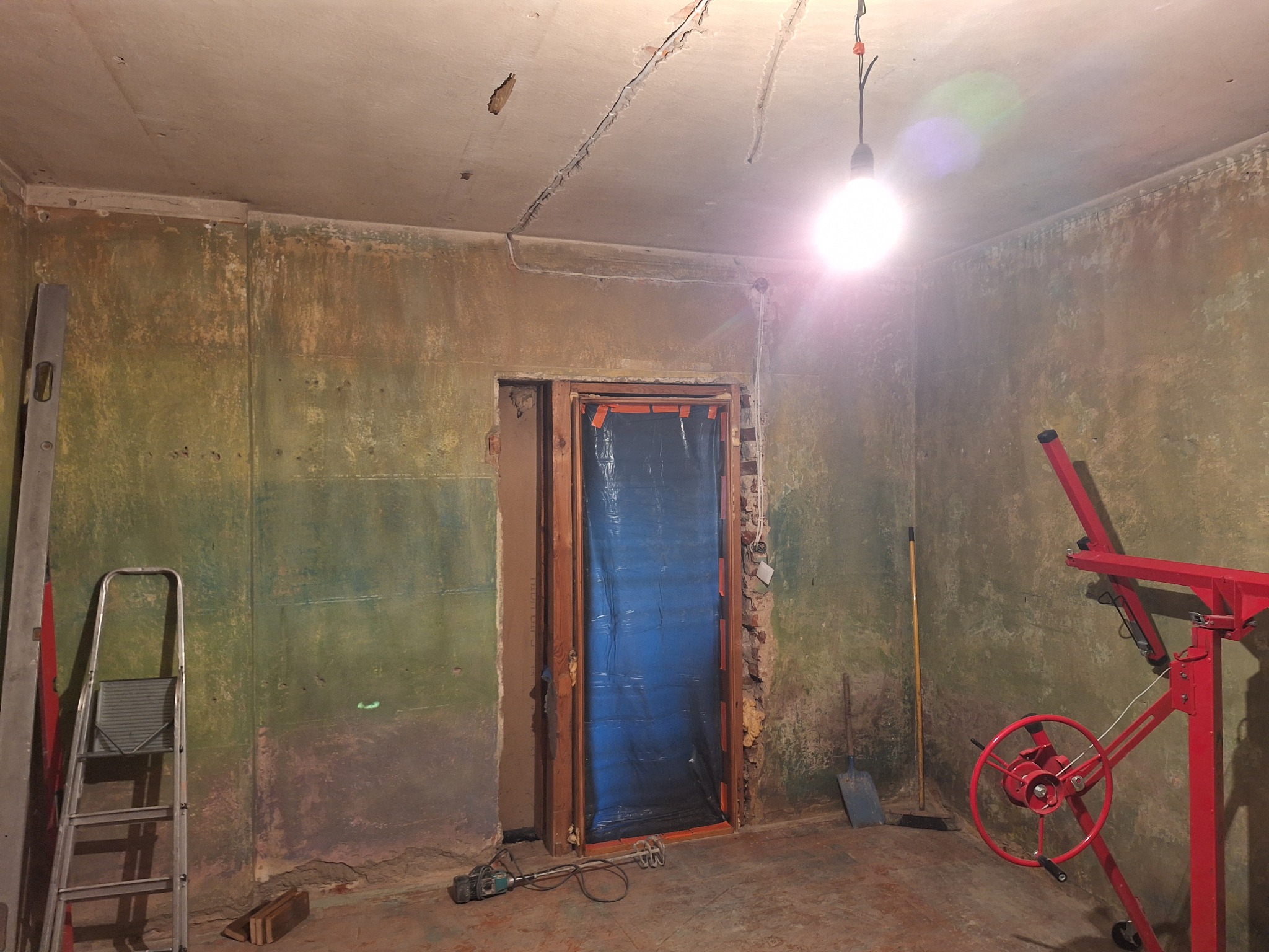 Pokój w trakcie remontu: ściany z resztkami farby, zabezpieczone drzwi, drabina, poziomica i podnośnik do płyt gipsowych. Widoczne ślady po instalacjach na suficie.