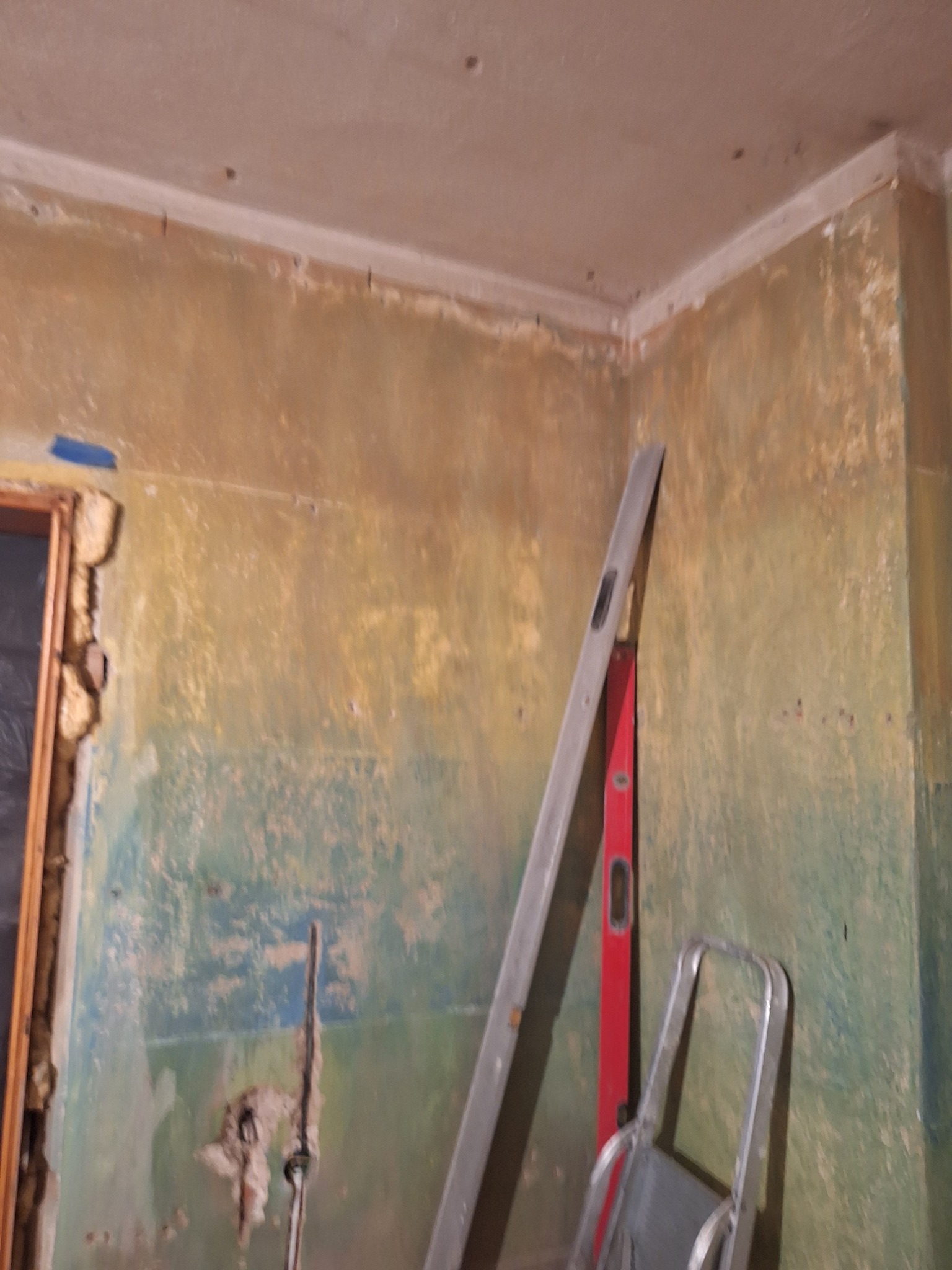 Przygotowanie ścian do prac wykończeniowych: surowe ściany z pozostałościami farby, drabina oparta o ścianę, poziomica. Widoczne ślady po usuniętych elementach.