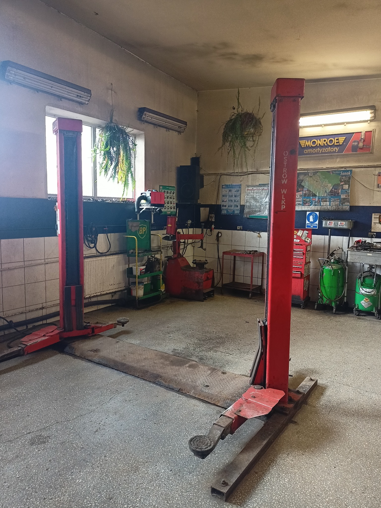 Wnętrze warsztatu samochodowego z podnośnikiem dwukolumnowym, narzędziami i roślinami doniczkowymi. Widoczne plakaty i akcesoria warsztatowe.