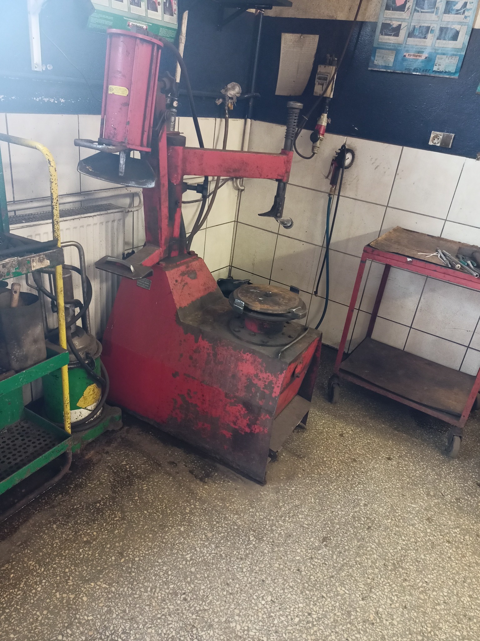 Czerwona, używana montażownica do opon w warsztacie. Obok wózek narzędziowy i stolik warsztatowy. Widoczne płytki na ścianie i betonowa posadzka.