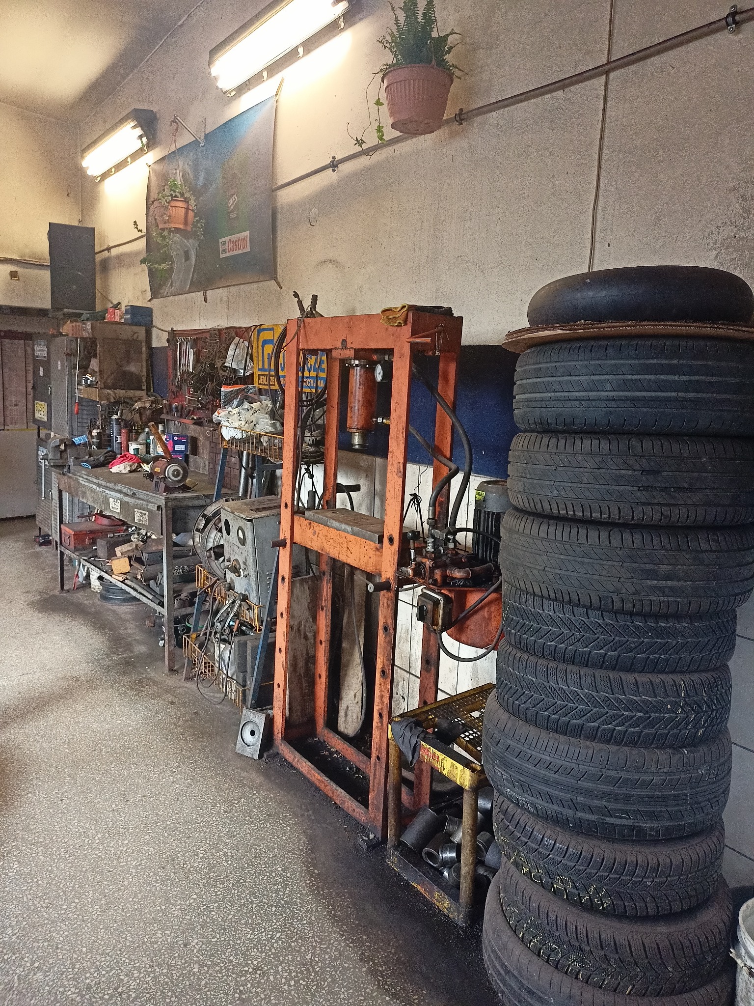 Wnętrze warsztatu: stos opon obok pomarańczowej prasy hydraulicznej, stół warsztatowy z narzędziami, plakat Castrol i doniczka z paprotką pod sufitem.