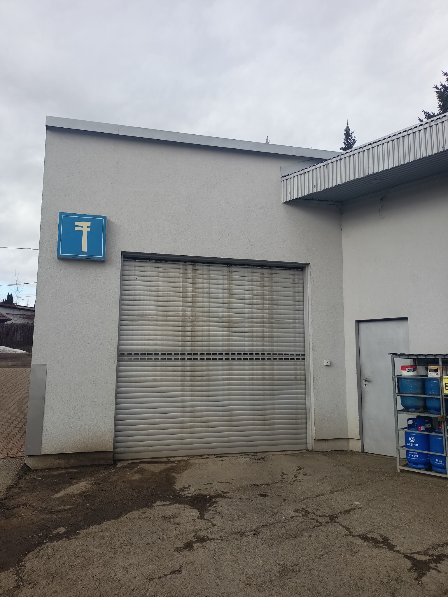 Szary budynek warsztatu z niebieskim znakiem z symbolem klucza francuskiego nad brudną, metalową bramą rolowaną i regałem z butlami gazowymi obok drzwi.