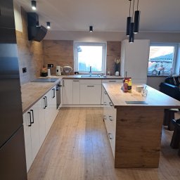 dan-pol - Nowoczesna kuchnia z wyspą: białe szafki z czarnymi uchwytami, drewniany blat, podłoga laminowana. Minimalistyczne oświetlenie, okno z widokiem na zewnątrz.