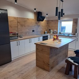 dan-pol - Nowoczesna kuchnia z wyspą i drewnianymi akcentami. Białe szafki kontrastują z drewnopodobną okładziną ściany. Czarny okap i lampy dodają elegancji.