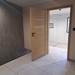 dan-pol - Otwarta, drewniana, nowoczesna  ościeżnica z przeszkleniami, prowadząca do wykończonej łazienki z marmurowymi płytkami. Widok z korytarza z szarą ścianą.