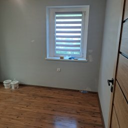dan-pol - Pokój po szpachlowaniu i malowaniu na szaro, widoczne dwa wiadra farby, drewniana podłoga i okno z żaluzjami. Świeżo pomalowane ściany, gotowe do dalszych prac wykończeniowych.