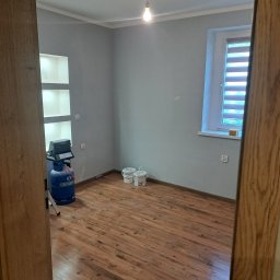 dan-pol - Jasnoszary pokój po malowaniu z drewnianą podłogą. Widoczne puszki z farbą, butla gazowa i wnęka w ścianie. Ujęcie zza framugi drzwi.