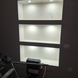 dan-pol - Ściana w odcieniach szarości z trzema wnękami oświetlonymi punktowo. Widoczny sprzęt malarski i drabina, sugerujące etap prac wykończeniowych.