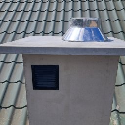 dan-pol - Wykończony komin z metalowym daszkiem i kratką wentylacyjną na tle dachu z szarej dachówki. Detal wykończenia komina, widok z bliska.