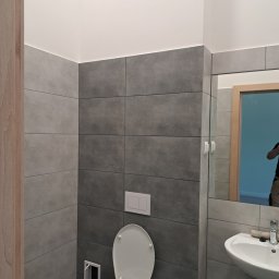 dan-pol - Nowoczesna łazienka z szarymi płytkami, białym sanitariatem i drewnianym akcentem ramy lustra. Minimalistyczny design, widoczny fragment osoby w odbiciu lustra.