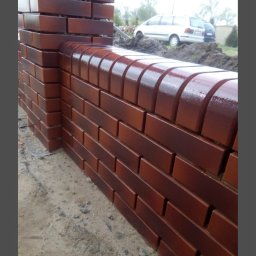 dan-pol - Błyszczący, brązowy mur z cegły klinkierowej, wykończony zaokrąglonymi elementami. W tle widoczny samochód i zieleń. Ujęcie z dołu, podkreślające strukturę cegieł.