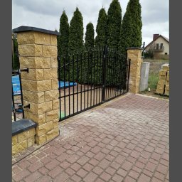 dan-pol - Nowa, czarna, metalowa brama wjazdowa z kamiennymi słupkami z piaskowca, prowadząca na posesję z kostką brukową. W tle tuje i dom.