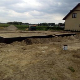 dan-pol - Fundamenty domu jednorodzinnego w trakcie budowy, widok z poziomu gruntu, z boku widoczny fragment budynku i sterta ziemi. Pochmurne niebo.