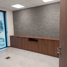 dan-pol - Biuro po remoncie: szara ściana z cegły dekoracyjnej, długa szafka z drewna, jasna podłoga z płytek. Widok na okno z żaluzjami i fragment drzwi z kluczem.