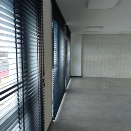 dan-pol - Nowoczesne, szare płytki podłogowe i ściana z białej cegły w jasnym wnętrzu z trzema oknami z czarnymi żaluzjami. Widok z boku, oświetlenie sufitowe.