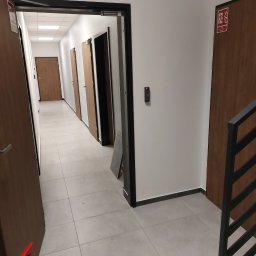 dan-pol - Korytarz biurowy z szarymi płytkami, brązowymi drzwiami i systemem kontroli dostępu. Widoczny znak ewakuacyjny i gaśnica. Oświetlenie sufitowe.
