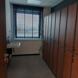 dan-pol - Wnętrze szatni z umywalką i rzędem szafek zamykanych na klucz. Widok na okno z żaluzjami i sufit z kasetonami. Minimalistyczny design.