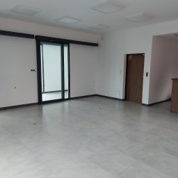 dan-pol - Jasne, przestronne wnętrze biurowe z szarą podłogą z płytek, białymi ścianami i sufitowymi lampami LED. Widoczne przesuwne drzwi i drzwi przeciwpożarowe.