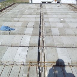 dan-pol - Wieniec stropowy zbrojony stalą, wypełniony betonowymi bloczkami szalunkowymi. Na bloczkach leży niebieska szmatka. Widoczny cień osoby robiącej zdjęcie.