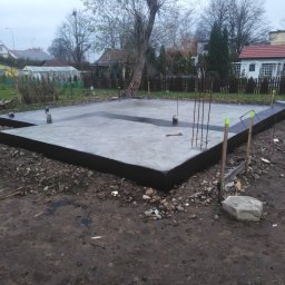 dan-pol - Wylana płyta fundamentowa z izolacją pionową, wystające pręty zbrojeniowe, teren budowy w tle zabudowa jednorodzinna, pochmurny dzień.