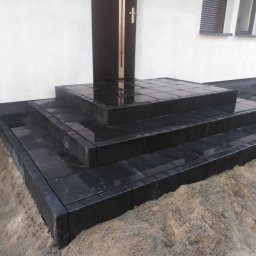 dan-pol - Nowoczesne, czarne schody wejściowe z kostki brukowej, prowadzące do ciemnych drzwi. Widoczne okno z żaluzjami. Otoczenie w trakcie prac wykończeniowych.