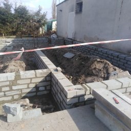 dan-pol - Fundamenty domu w trakcie budowy: ściany z bloczków betonowych, wykop, taśma ostrzegawcza. Widoczne narzędzia murarskie i fragment budynku w tle.