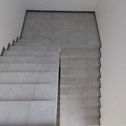 dan-pol - Wykończone schody wewnętrzne z jasnych, kwadratowych płytek, widok z góry. Minimalistyczny design, białe ściany, ciemna listwa przy podłodze.