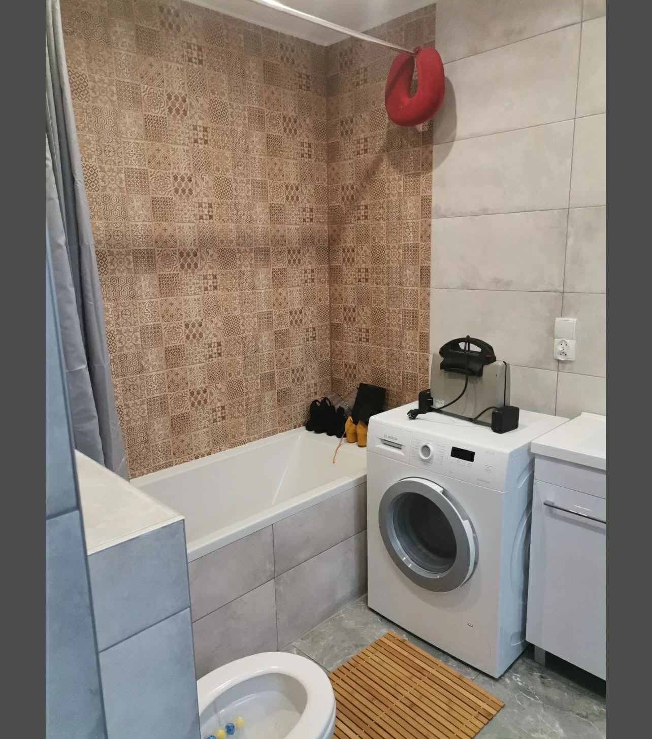 Ukończona łazienka z brązowymi płytkami wzorzystymi na ścianie, białą wanną i pralką. Widoczny fragment toalety i bambusowa mata podłogowa.