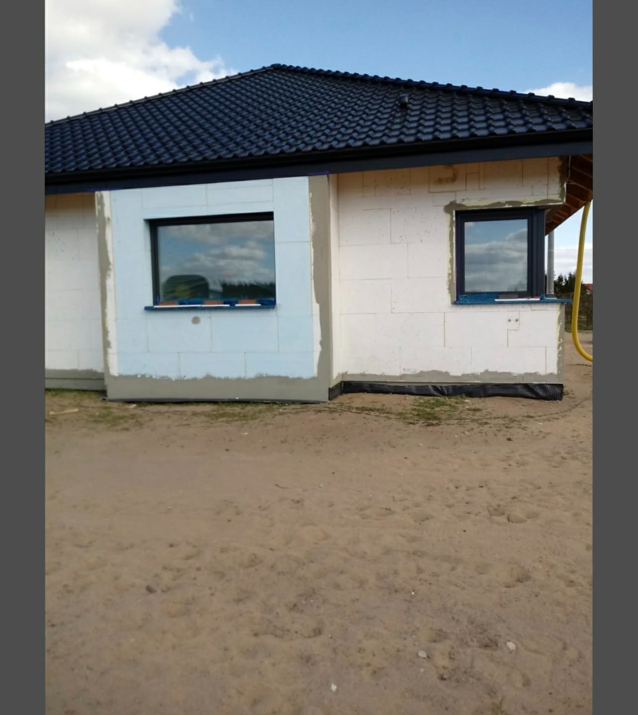 Niedokończony dom jednorodzinny z białego bloczka, z czarną dachówką i oknami. Widoczne ocieplenie i przygotowanie do dalszych prac wykończeniowych.