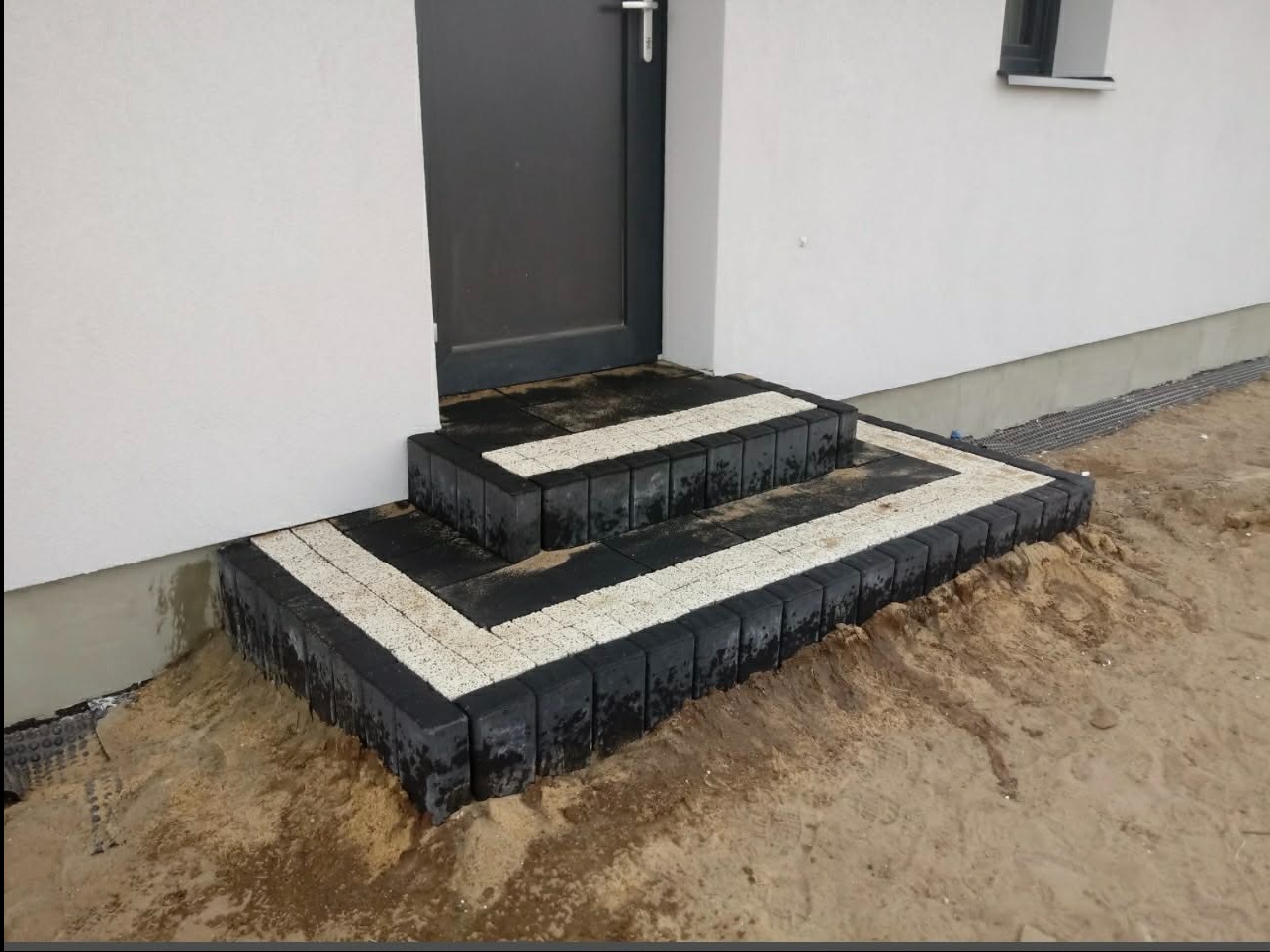 Schody wejściowe z kostki brukowej w odcieniach szarości i bieli, kontrastujące z jasną elewacją budynku. Widoczne detale ułożenia kostki i surowe otoczenie.