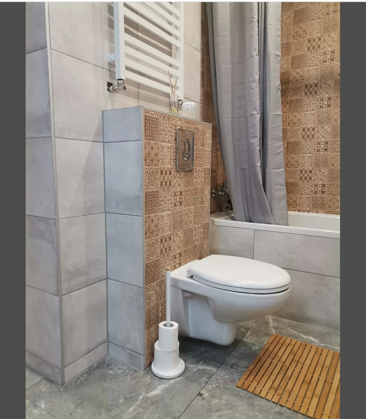 Wykończona łazienka z podwieszaną toaletą, brązowo-beżowymi płytkami z wzorem, szarą zasłoną prysznicową i grzejnikiem drabinkowym. Na podłodze mata bambusowa.