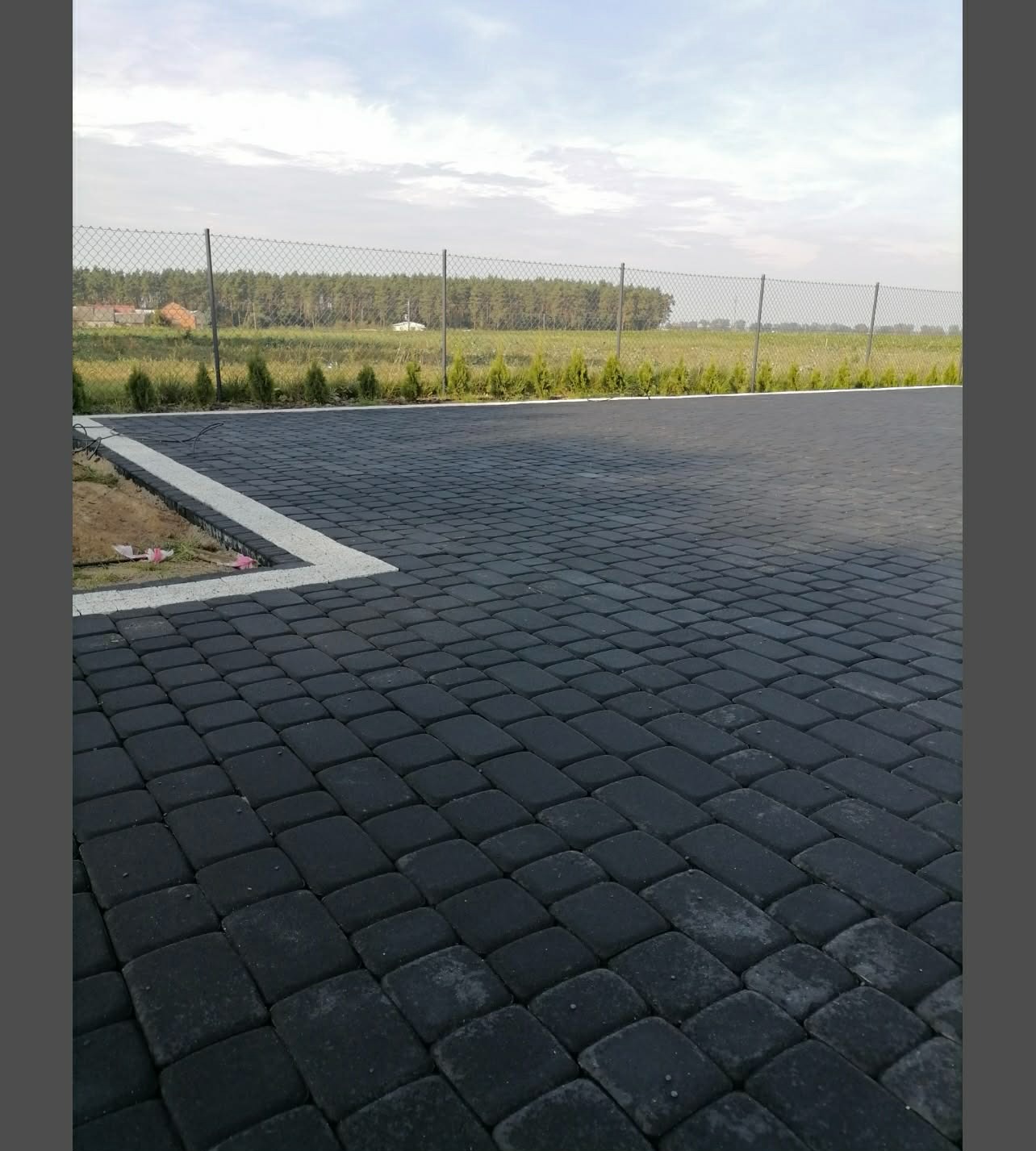 Ułożona, czarna kostka brukowa z białym obramowaniem na posesji. W tle siatka ogrodzeniowa, zieleń i las. Widok z perspektywy ziemi, w pochmurny dzień.