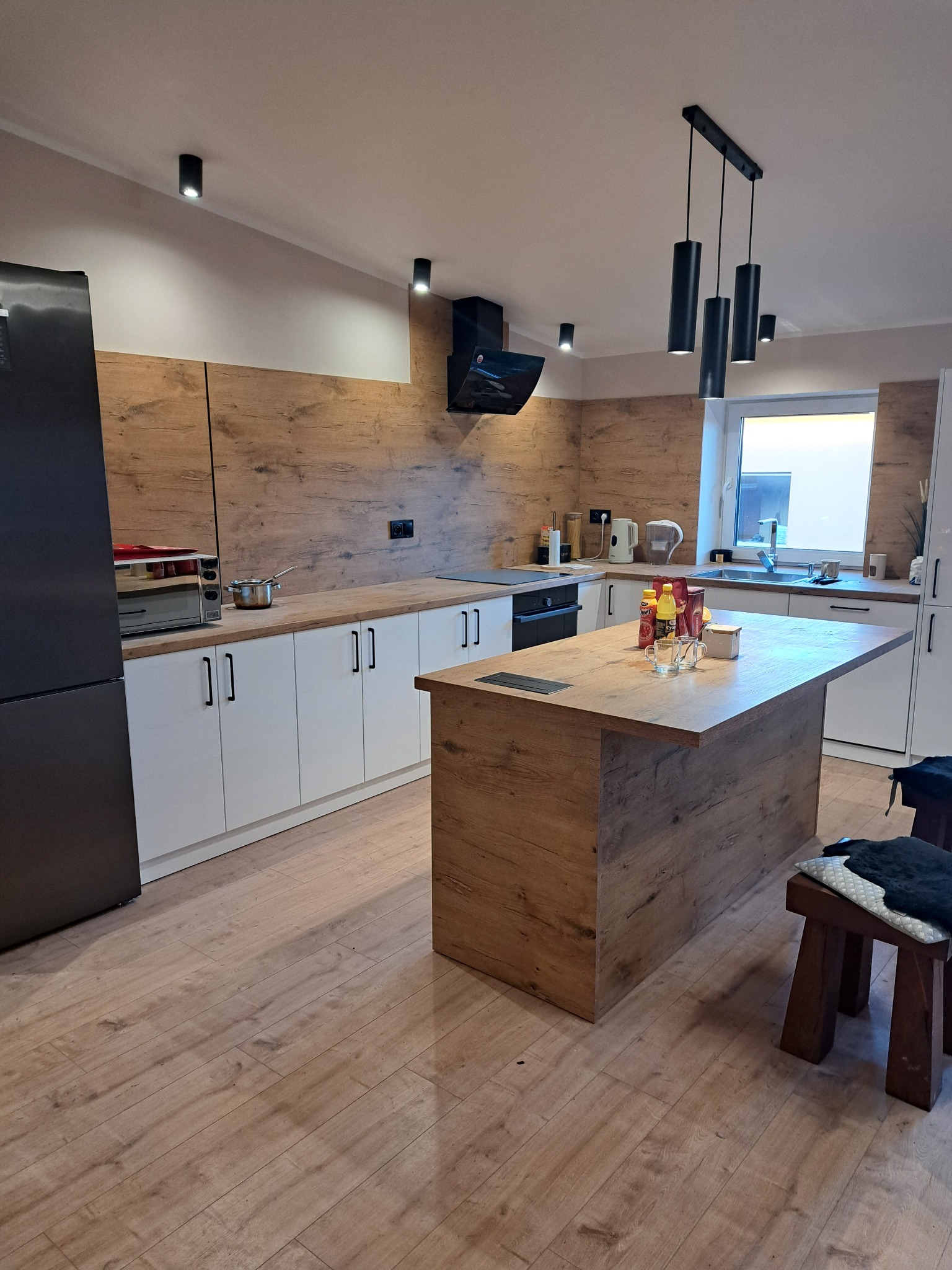 Nowoczesna kuchnia z wyspą i drewnianymi akcentami. Białe szafki kontrastują z drewnopodobną okładziną ściany. Czarny okap i lampy dodają elegancji.