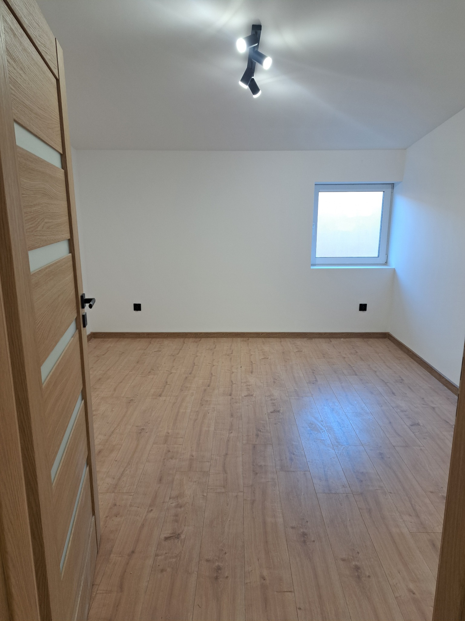 Jasne wnętrze po remoncie: drewniana podłoga, białe ściany, okno i nowoczesne oświetlenie sufitowe. Otwarta drewniana  ościeżnica drzwi z lewej strony kadru.