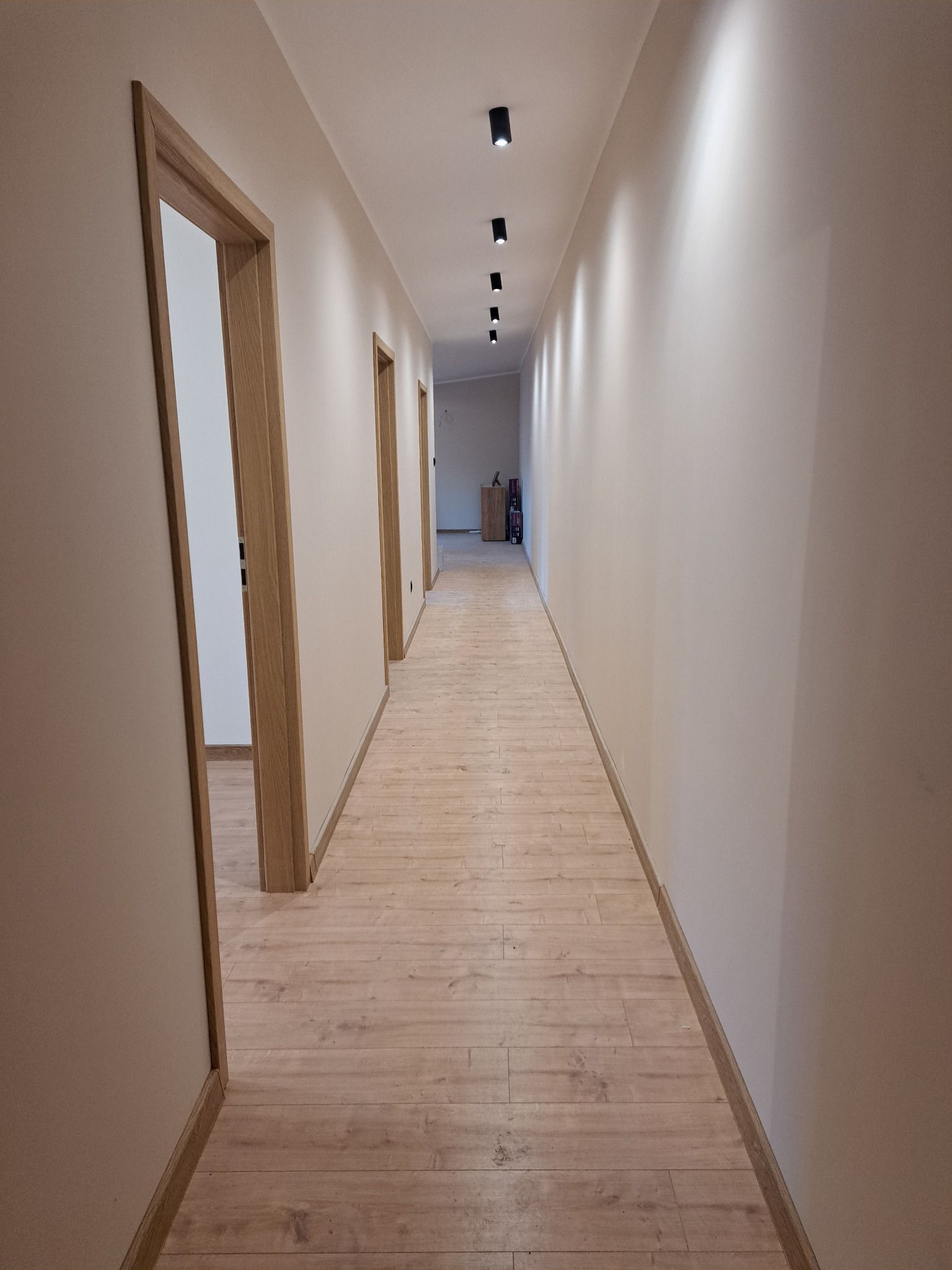 Długi korytarz po remoncie z drewnianą podłogą i ościeżnicami. Ściany w odcieniach beżu, oświetlenie punktowe sufitowe. Widok w głąb, perspektywa zbieżna.