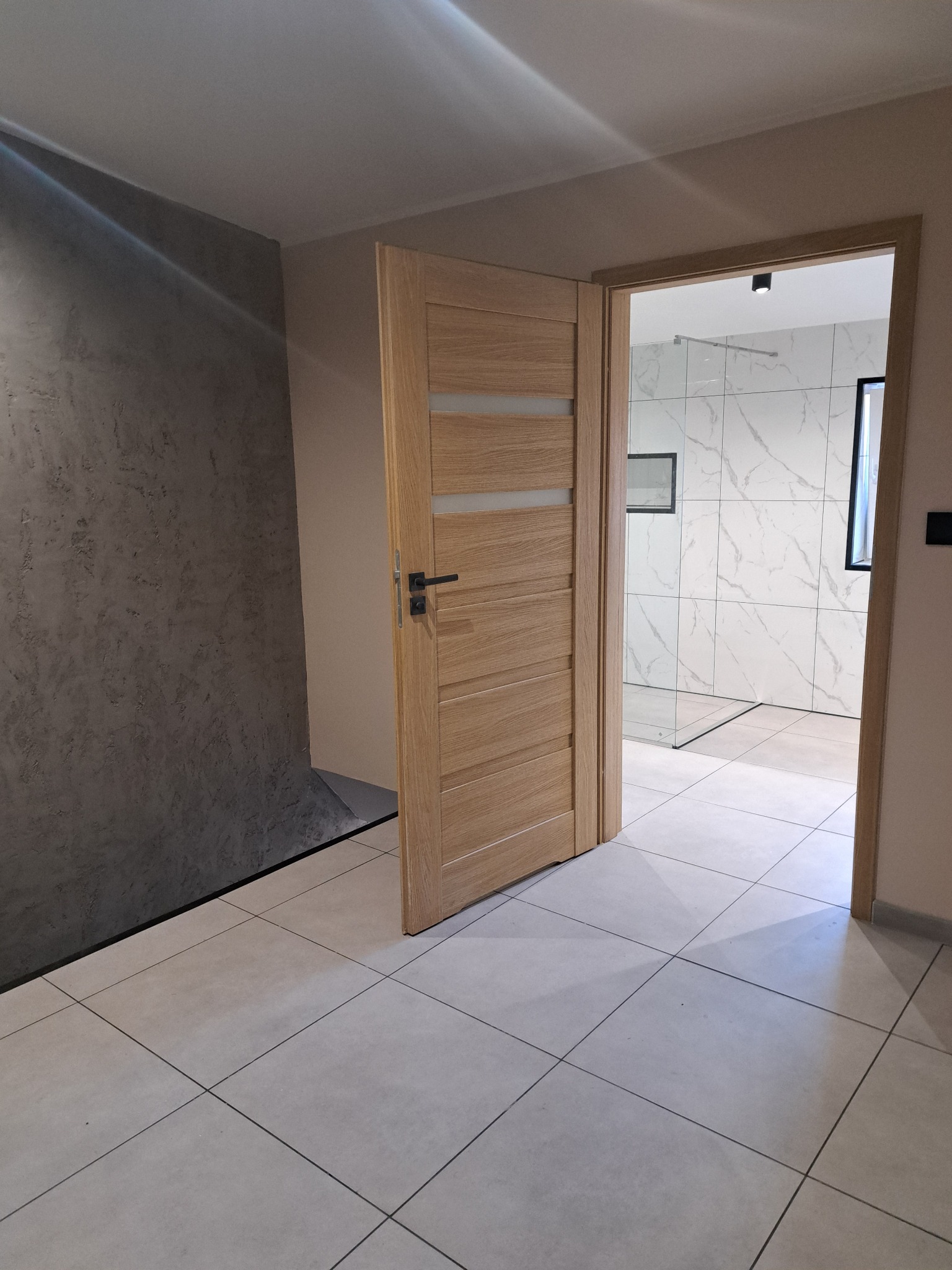 Otwarta, drewniana, nowoczesna  ościeżnica z przeszkleniami, prowadząca do wykończonej łazienki z marmurowymi płytkami. Widok z korytarza z szarą ścianą.