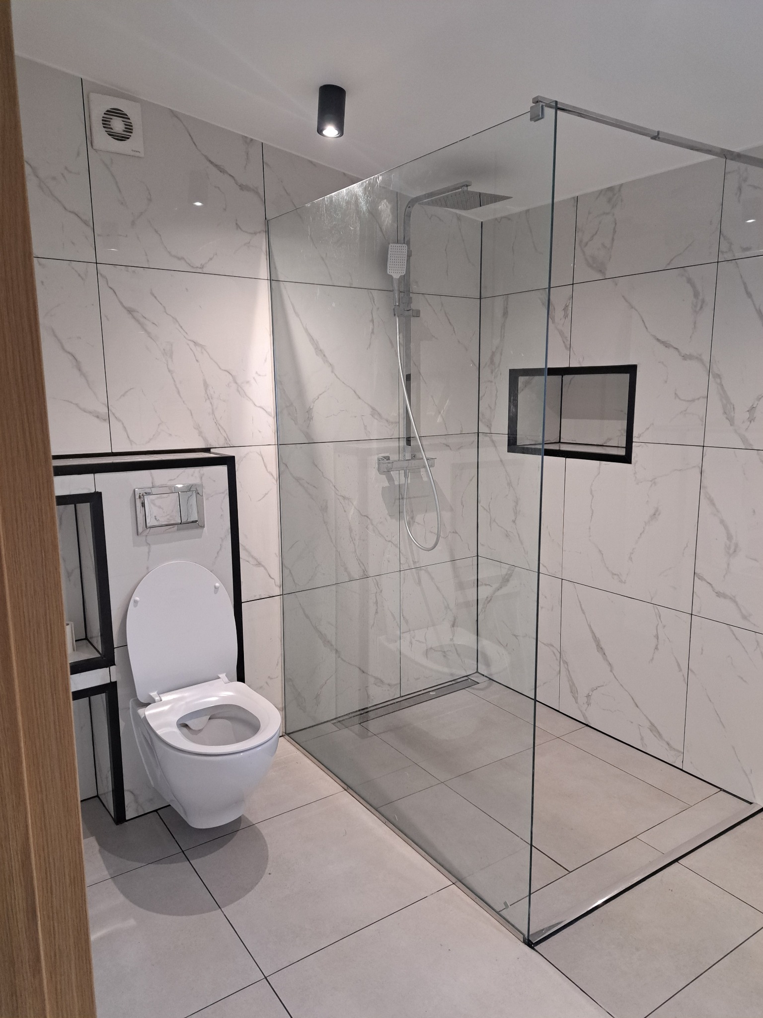 Nowoczesna łazienka z kabiną typu walk-in i podwieszaną toaletą. Białe płytki imitujące marmur z czarnymi akcentami. Minimalistyczny design, szklana ściana kabiny.