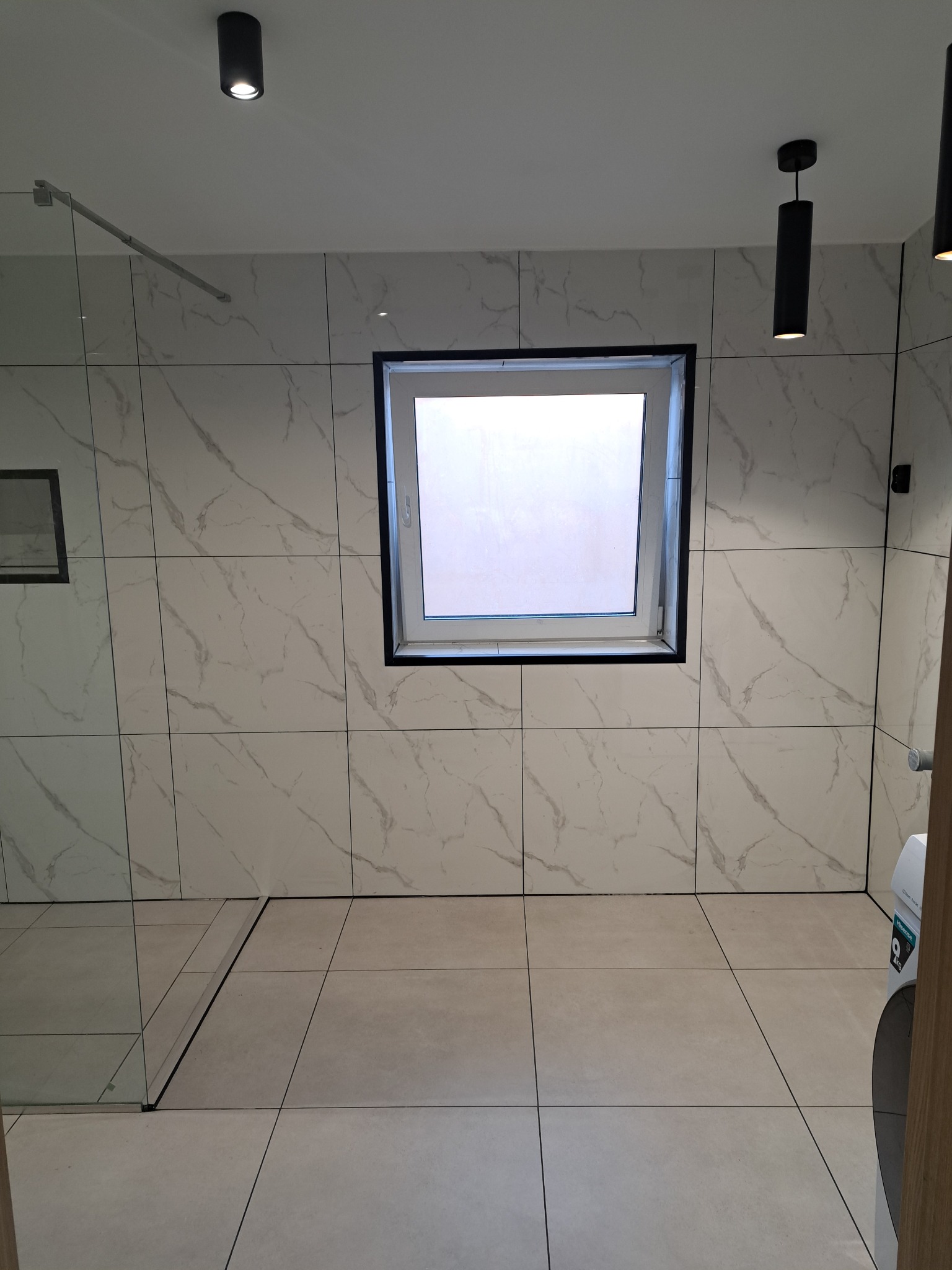 Wykończona łazienka z kabiną prysznicową typu walk-in, płytkami imitującymi marmur na ścianach i beżowymi płytkami na podłodze. Widoczne czarne, minimalistyczne lampy.