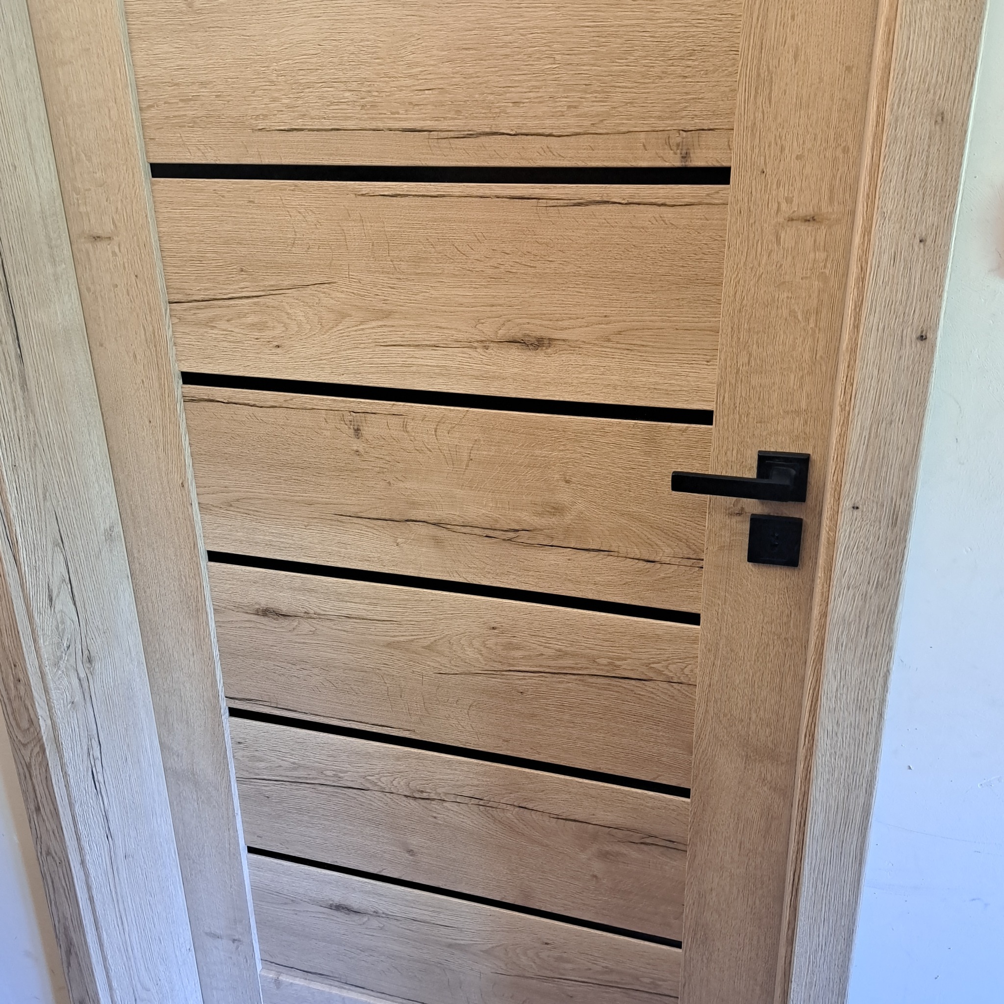 Drewniane drzwi wewnętrzne z poziomymi, czarnymi wstawkami i czarną klamką, ukazane z bliska. Widoczna faktura drewna i minimalistyczny design.