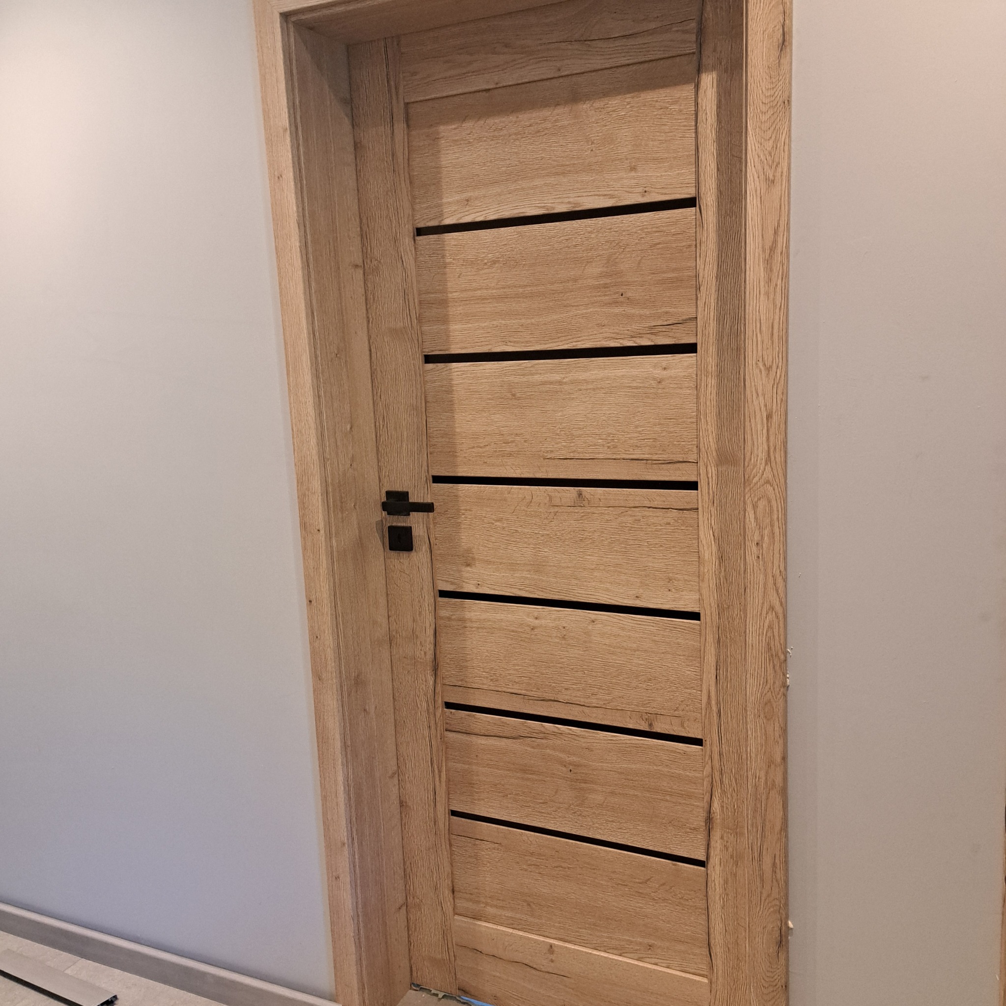 Nowoczesne, drewniane drzwi wewnętrzne z poziomymi, czarnymi wstawkami i czarną klamką, zamontowane w jasnym pomieszczeniu. Widoczna faktura drewna.