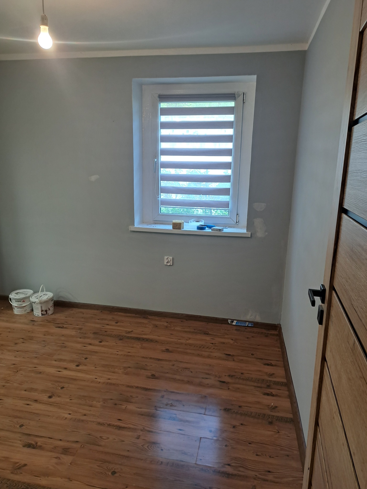 Pokój po szpachlowaniu i malowaniu na szaro, widoczne dwa wiadra farby, drewniana podłoga i okno z żaluzjami. Świeżo pomalowane ściany, gotowe do dalszych prac wykończeniowych.