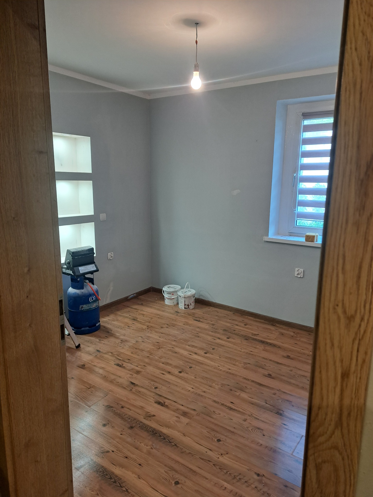 Jasnoszary pokój po malowaniu z drewnianą podłogą. Widoczne puszki z farbą, butla gazowa i wnęka w ścianie. Ujęcie zza framugi drzwi.